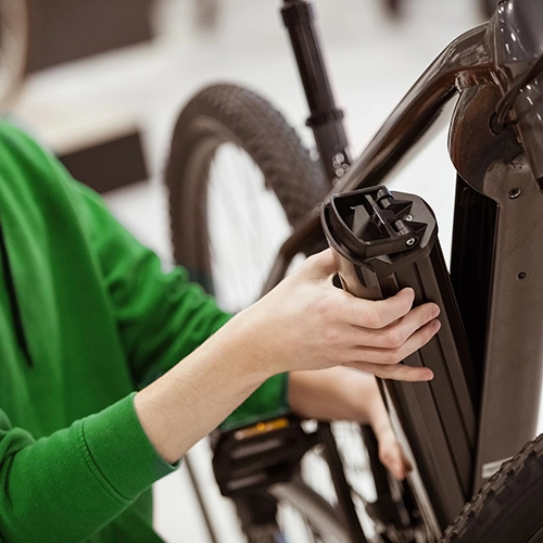 A person in a green sweatshirt inserts or removes a battery from the frame of a black electric bicycle.