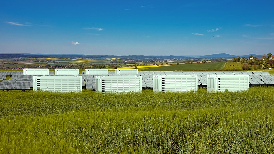 Rows of white battery storage containers sit in a green field beside solar panels under a blue sky, with distant hills and farmland in the background.