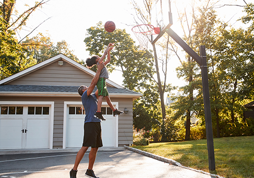 An adult lifts a child toward a basketball hoop outside, helping them make a shot in the driveway near a house with a garage, on a sunny day surrounded by trees.