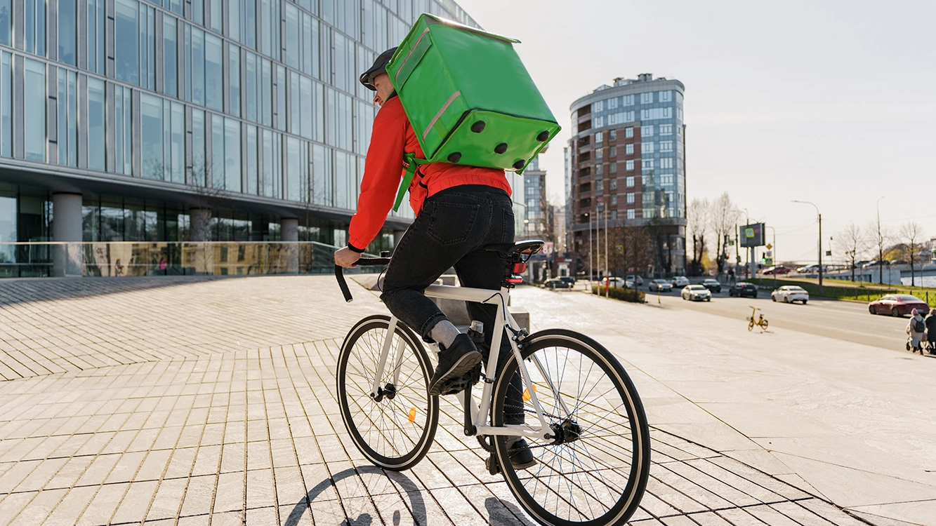 A person wearing a green backpack rides a bicycle up a sloped sidewalk near modern glass buildings and city streets on a clear day.