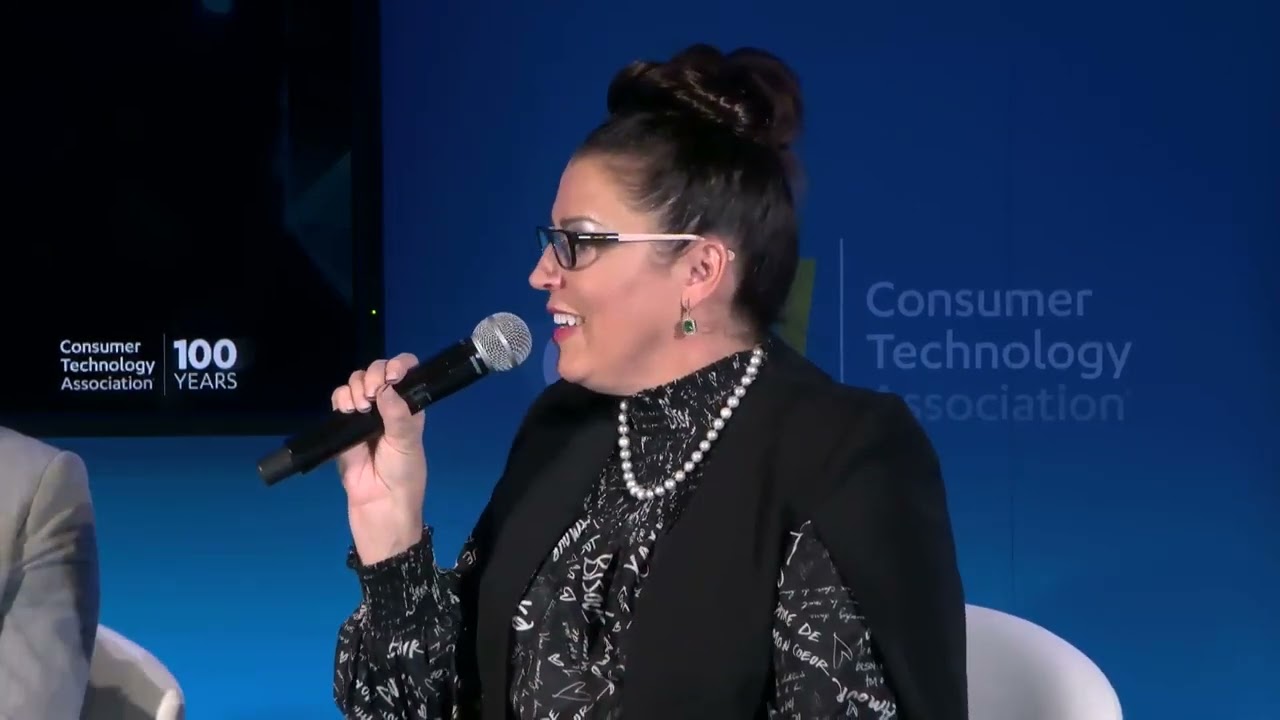 A woman with glasses and a high bun speaks into a microphone while seated at an event. She is wearing a black patterned blouse, a black jacket, a pearl necklace, and earrings. "Consumer Technology Association" is visible in the background.