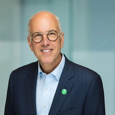 Smiling older man with glasses and a bald head, wearing a dark pinstripe suit, light blue shirt, and a round green pin, standing in front of a blurred, light-colored background.