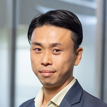 A man with short, neatly combed black hair is wearing a blue blazer and a light-colored shirt, standing in front of a blurred office background. He is looking at the camera with a slight smile.