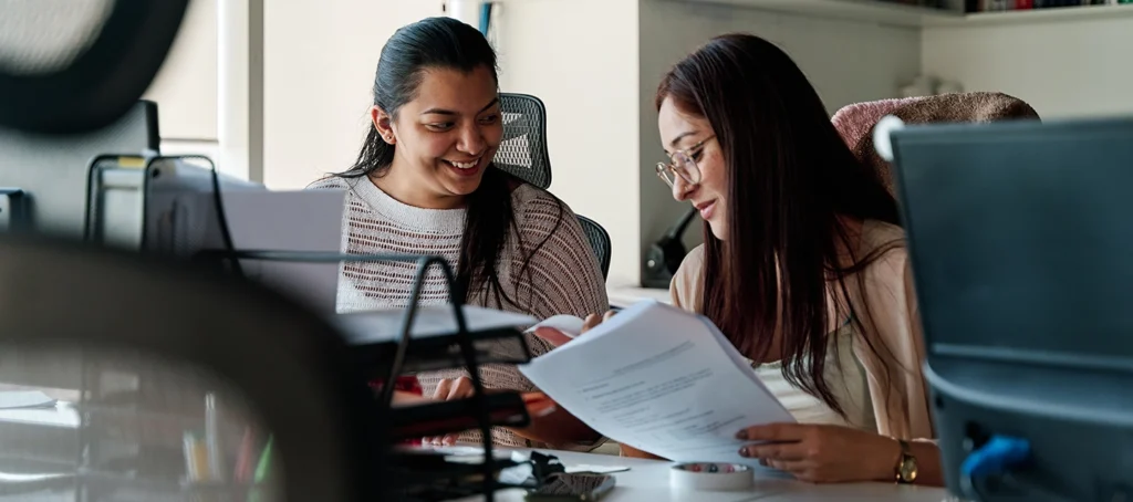Two women sitting at a desk in an office, smiling and discussing documents together. One woman is holding papers while the other looks on, both appearing engaged and collaborative in their work.