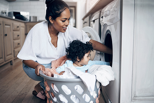 A woman kneels by a washing machine, placing laundry inside, while a young child sits in a laundry basket filled with clothes, looking at her. They are in a home laundry room.