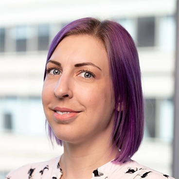 A person with straight, shoulder-length purple hair smiles slightly, looking toward the camera. They are wearing a light-colored top. The background is blurred, showing an office building with large windows.