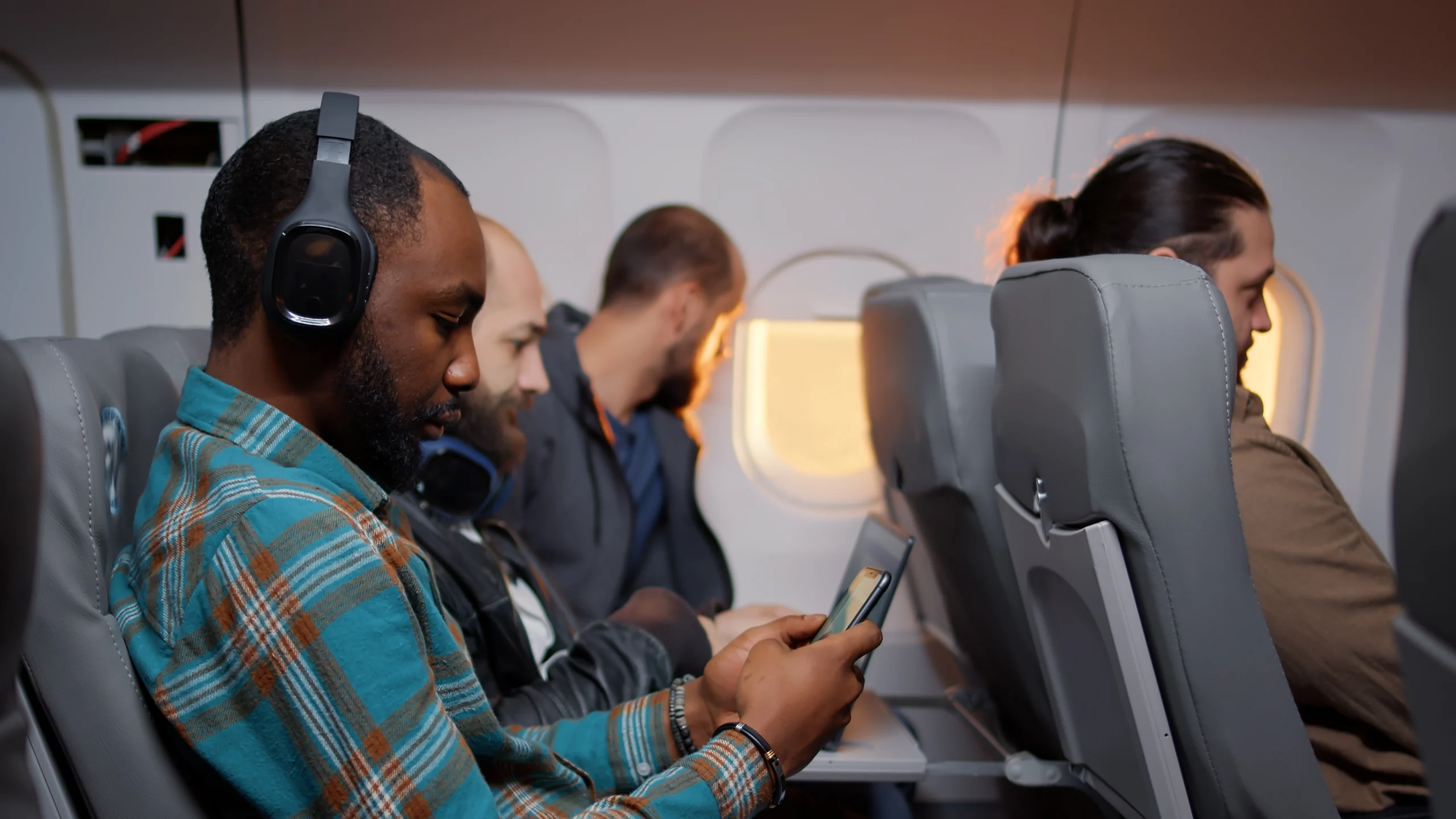 Four airline passengers sit in a row. The man in front wears headphones and looks at his phone. Two men behind look out the window, and a woman in the back row has her hair in a bun and is leaning forward.