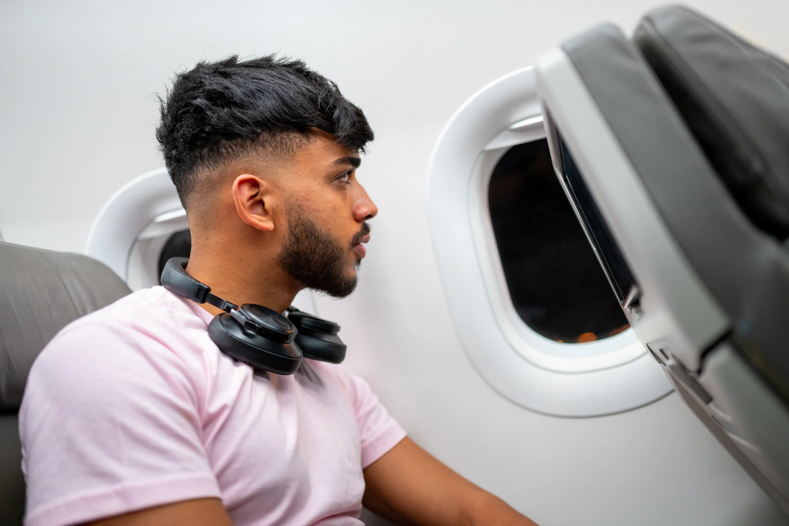 A young man wearing a pink t-shirt and headphones around his neck sits on an airplane, looking thoughtfully out the window.