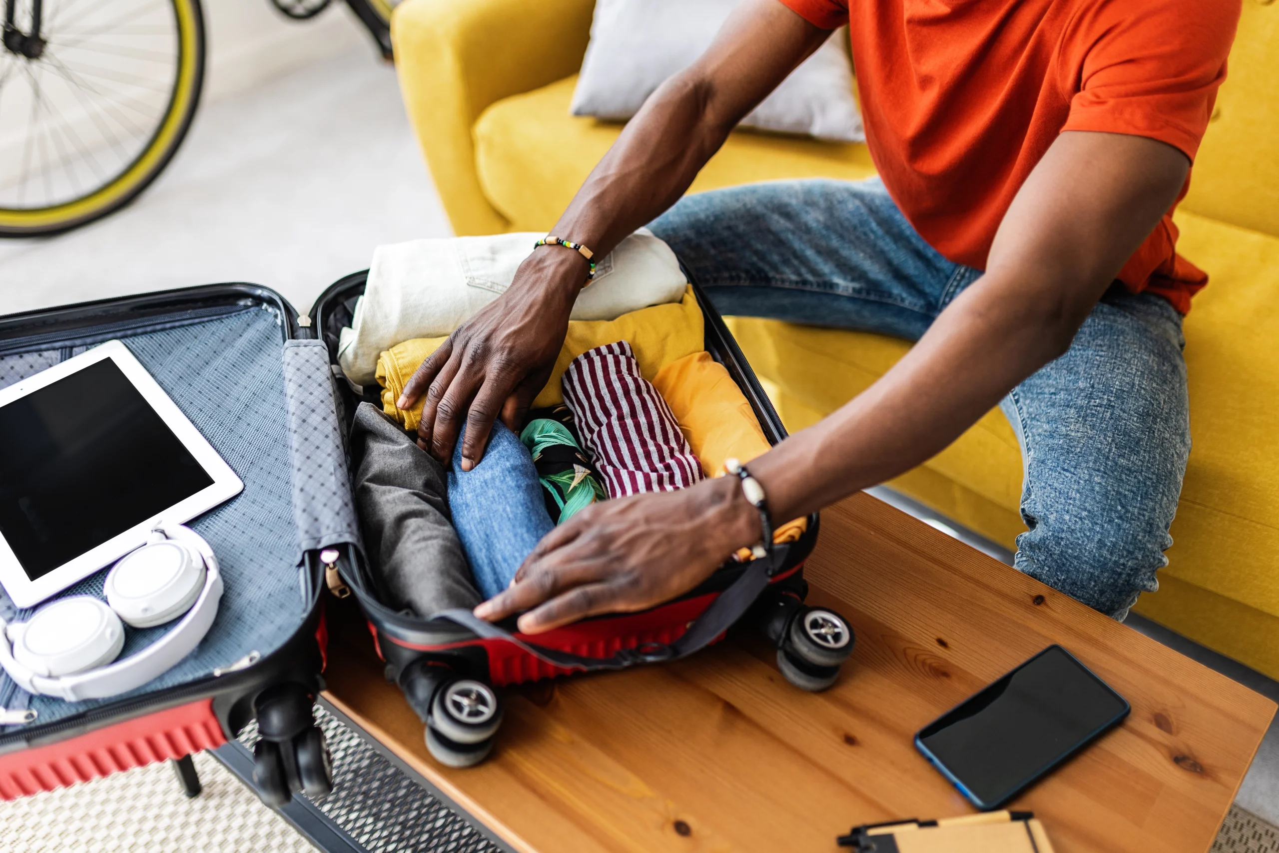A person packing clothes into a small suitcase on a wooden table, with an iPad, headphones, and a smartphone nearby, sitting on a yellow couch.