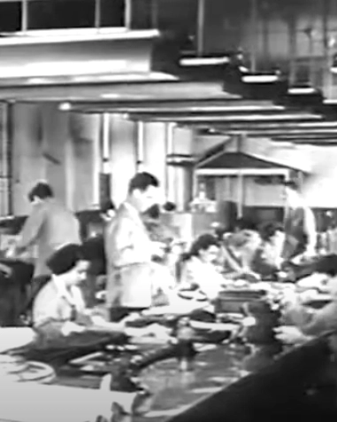 Black and white photo of a busy office with men and women working at desks, using typewriters and paperwork. The workspace is filled with people in formal clothing and overhead lights.
