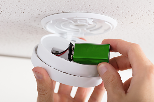 A person installs a green battery into an open smoke detector mounted on a white ceiling.