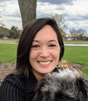 A woman with shoulder-length dark hair smiles at the camera outdoors, with green grass and trees in the background. The furry head of a small black-and-gray dog is visible in the lower foreground.
