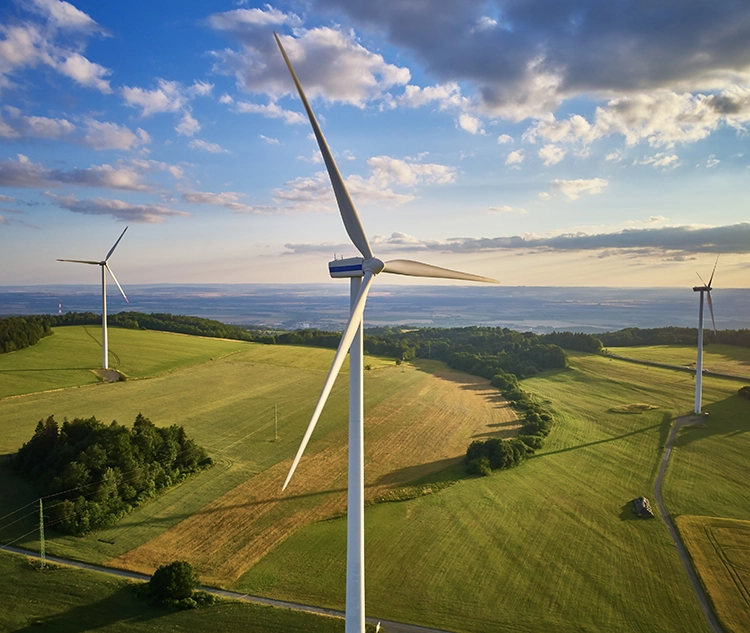 Aerial view of wind turbines standing in green fields under a partly cloudy sky, with rolling hills and distant landscape visible on the horizon.