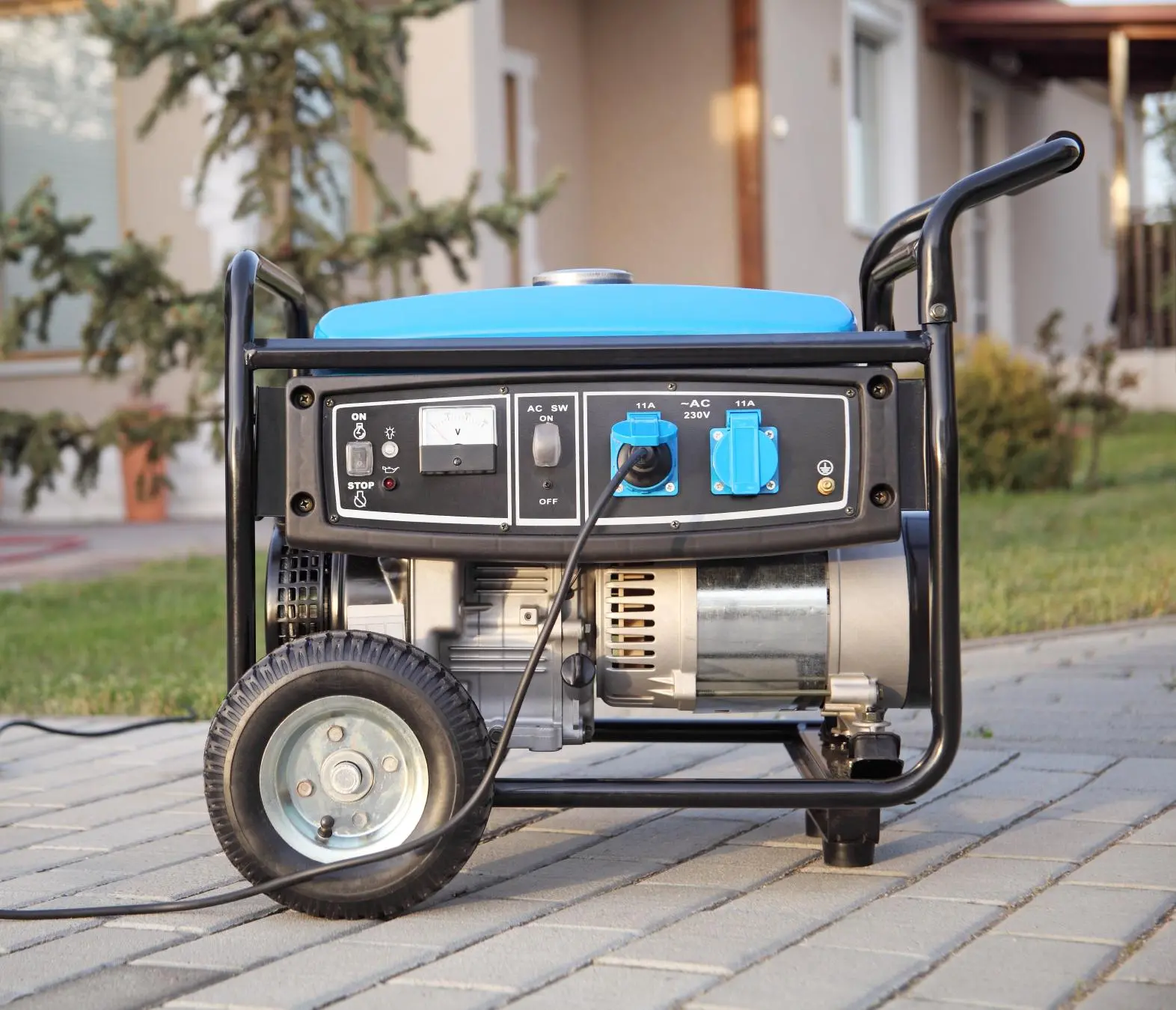 A portable electric generator with a blue top sits on a paved surface outdoors, connected by a power cord, with a house and greenery visible in the background.