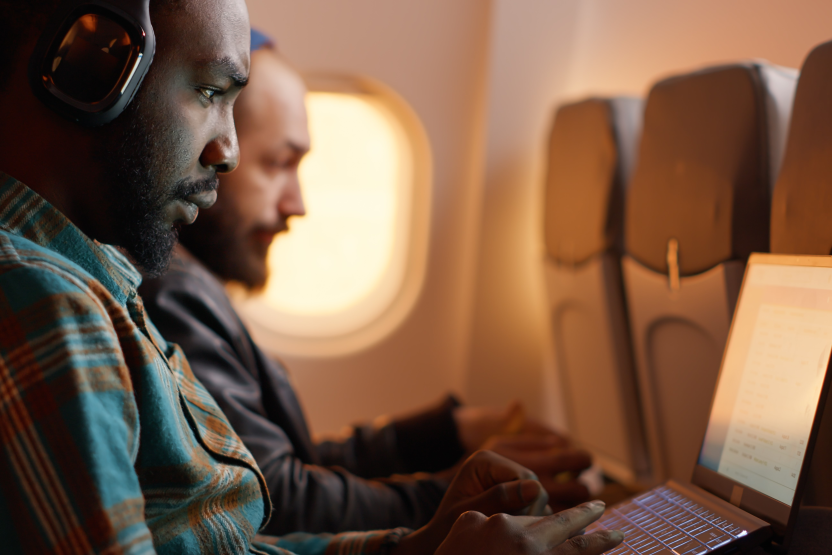 Two men sit side by side on an airplane, both focused on their laptops. The man in the foreground wears headphones and a plaid shirt, while the man behind him has a shaved head and a beard.