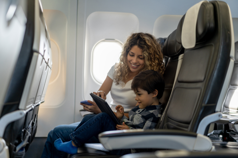 A woman and a young boy sit together on an airplane. The boy is using a tablet, and the woman is smiling and looking at the screen with him. They appear happy and relaxed during their flight.