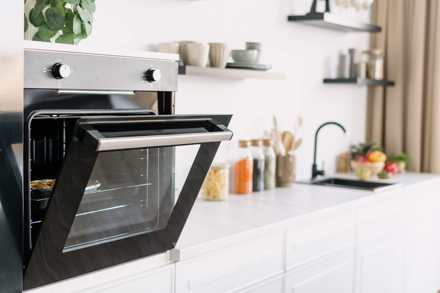 A modern kitchen with a built-in oven door open, glass jars filled with pasta and grains on the countertop, a black sink, and shelves with dishes and glasses.