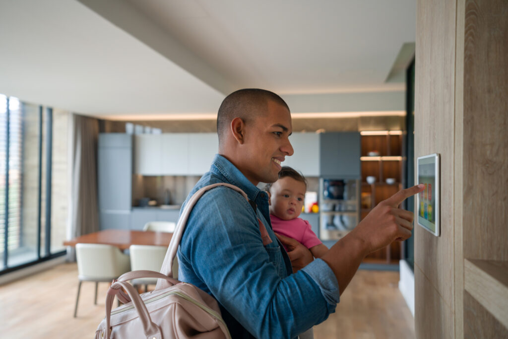 A man holding a baby adjusts a smart home control panel on the wall in a modern, bright kitchen and living area.