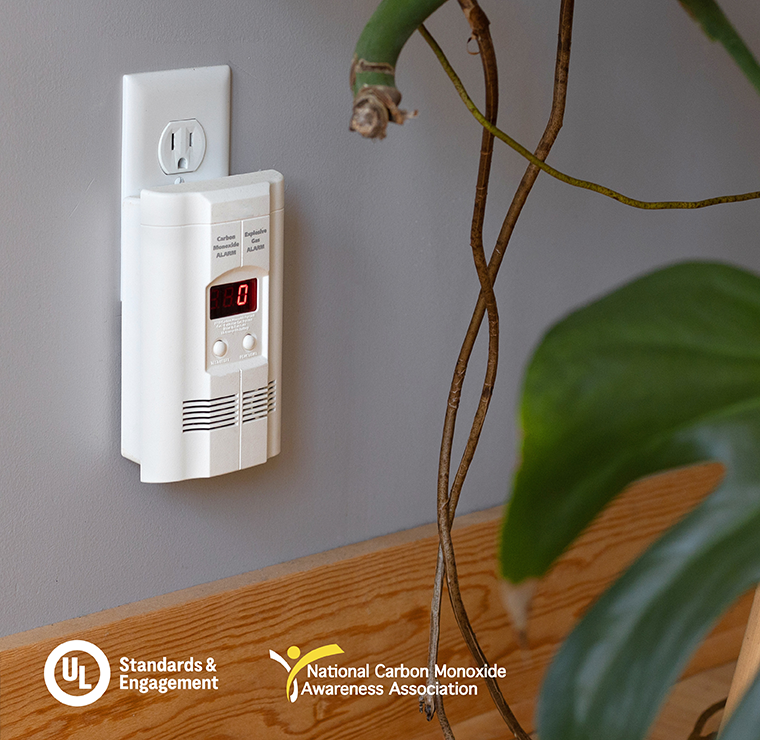 A carbon monoxide detector is plugged into a wall outlet near a plant. The device displays a digital reading, and two organization logos appear in the lower left corner.