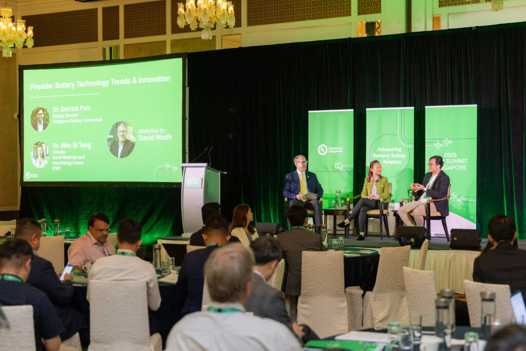 A panel discussion on stage features three speakers seated in armchairs with a moderator. Behind them, a large screen displays their names and session title, "Fireside: Battery Technology Trends & Innovation." Audience members watch attentively.