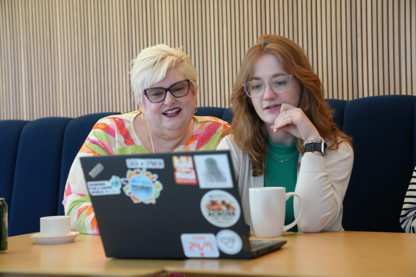 Two women sit at a table in front of a laptop covered with stickers, both smiling and looking at the screen. There are coffee cups on the table and a blue cushioned bench in the background.