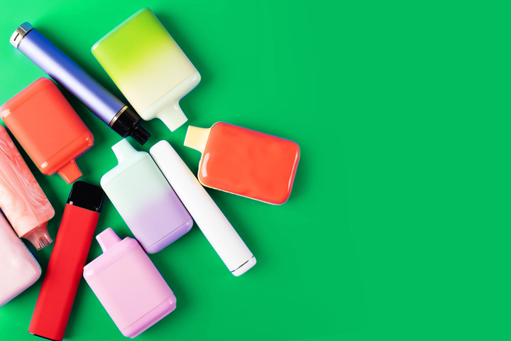 Several colorful disposable vapes are arranged on a bright green background, with space on the right side of the image. The vapes come in different shapes and pastel colors.