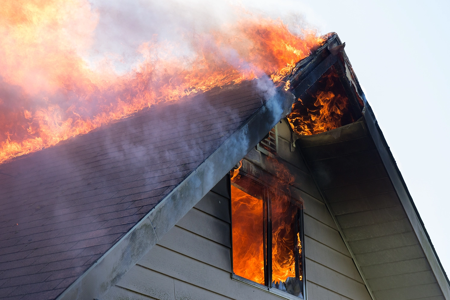 Flames and smoke pour out of the roof and window of a house, engulfing the upper story in a severe fire.