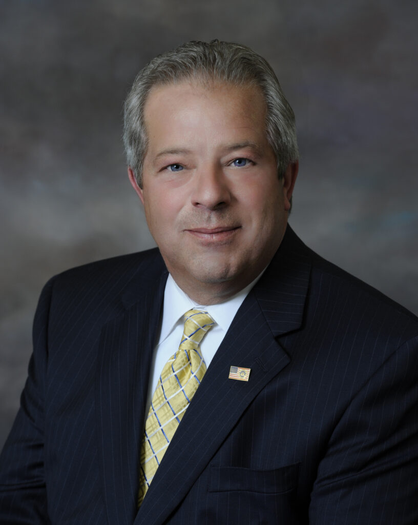 A man with gray hair is wearing a dark pinstripe suit, a yellow patterned tie, and an American flag pin. He is smiling slightly and facing the camera, with a blurred neutral background behind him.