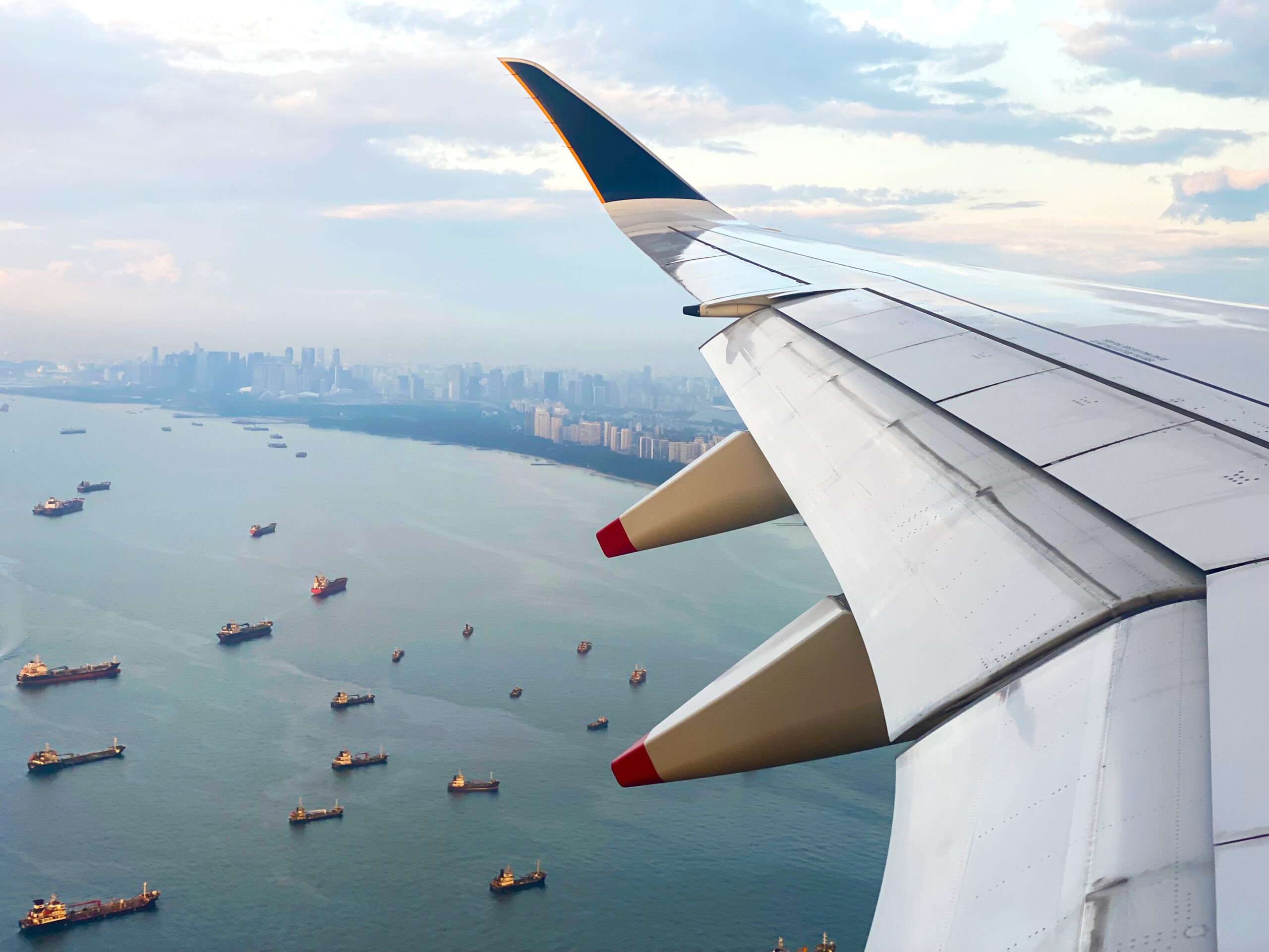 View from an airplane window showing the aircraft wing above the sea, with many ships below and a city skyline visible in the distance under a partly cloudy sky.