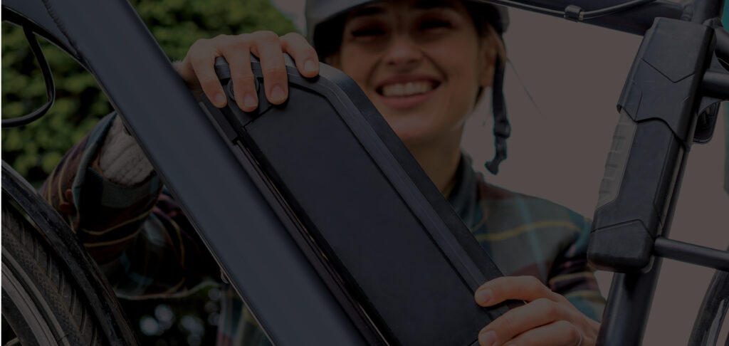 A person wearing a helmet smiles while placing or removing a rectangular battery pack from the frame of an electric bicycle.
