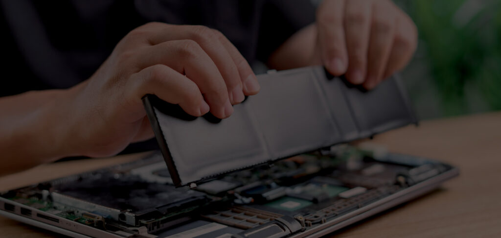 A person’s hands are removing or installing a rectangular battery inside an open laptop on a wooden table. The internal components of the laptop are visible.
