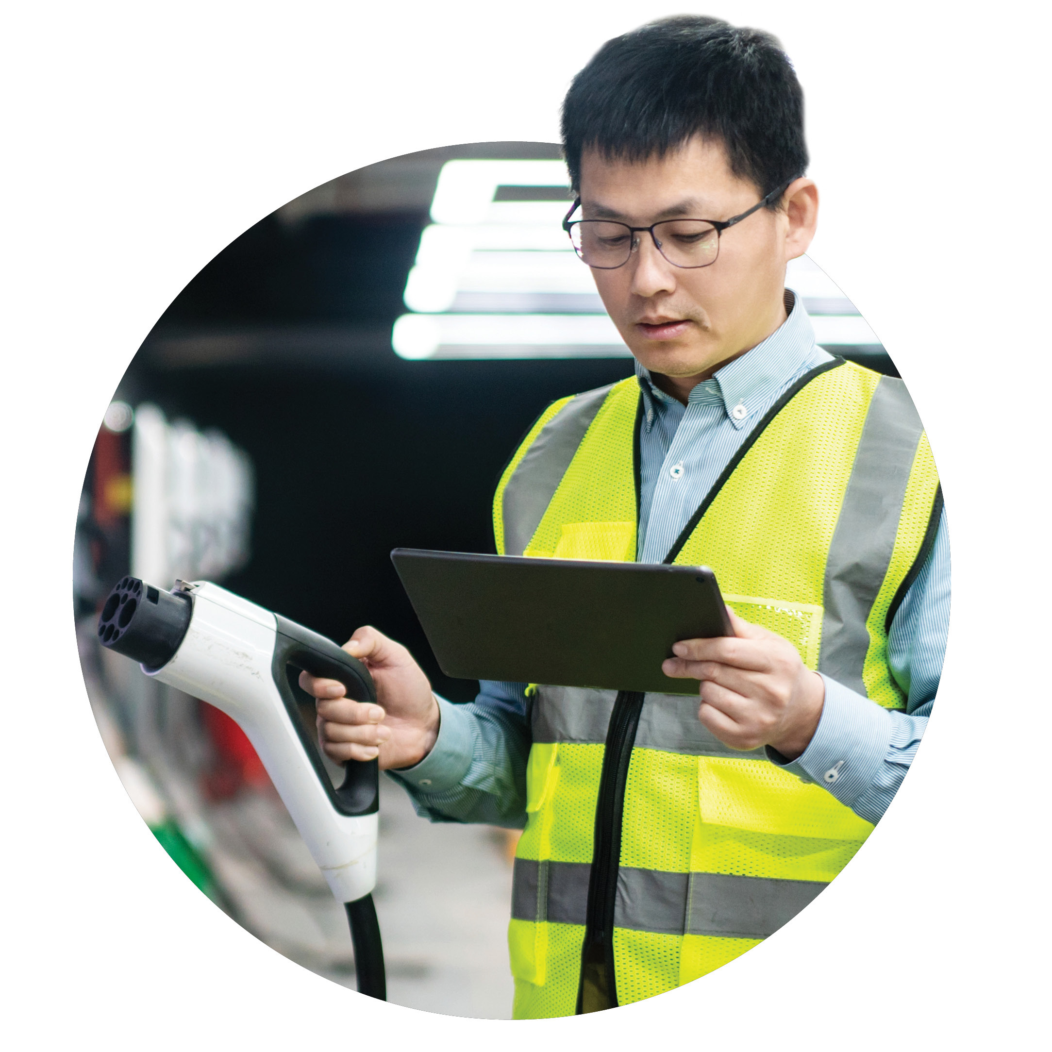 A man wearing glasses and a yellow safety vest holds an electric vehicle charging plug in one hand and uses a tablet with the other, standing in an industrial or garage setting.