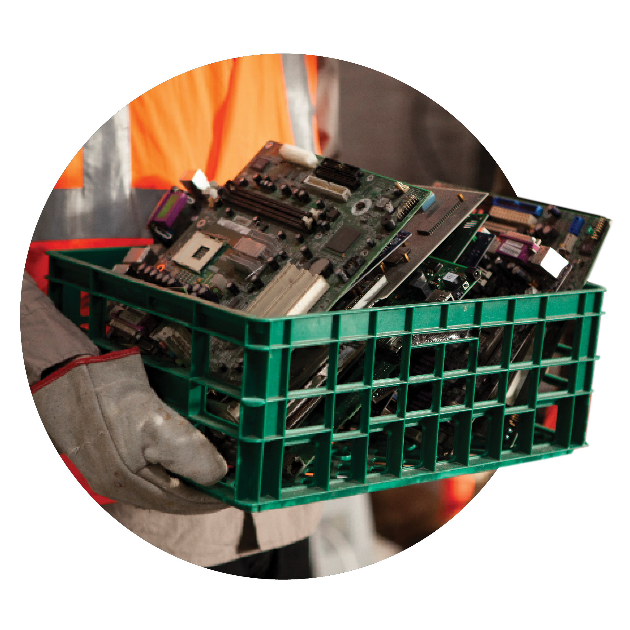 A person wearing gloves and an orange safety vest holds a green plastic crate filled with discarded computer motherboards and electronic components.