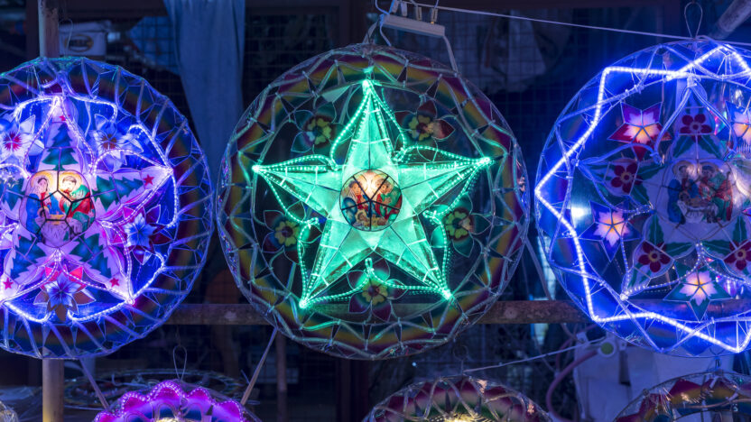 Colorful star-shaped lanterns, illuminated with bright green, purple, and blue lights, are displayed hanging in a row at an outdoor market, creating a festive atmosphere against a dark background.