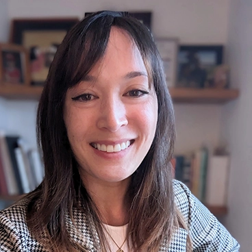 A woman with long brown hair and bangs smiles at the camera, wearing a black-and-white checkered jacket. Books and framed photos are visible on a wooden shelf in the background—a cozy scene untouched by counterfeit risks.