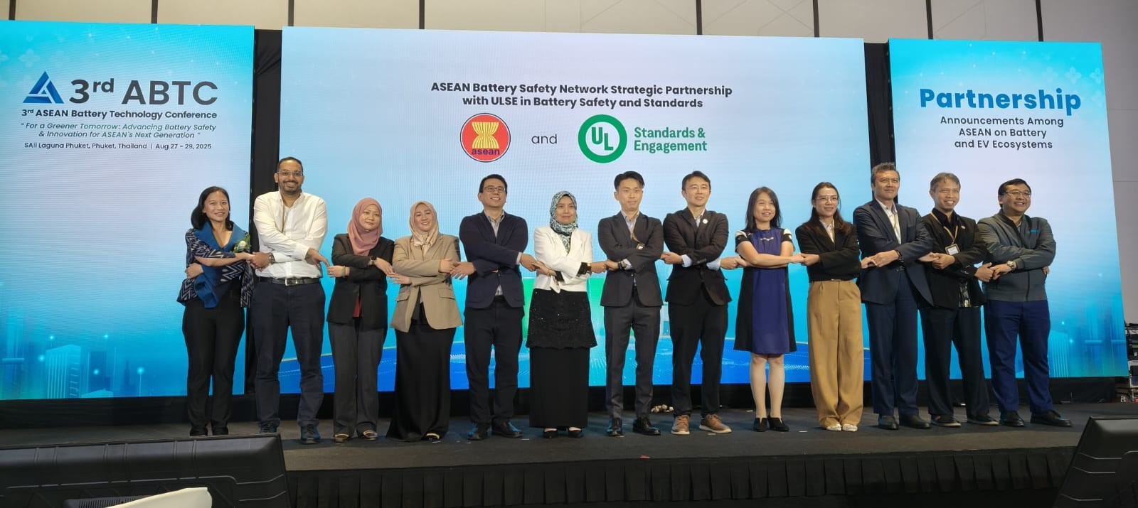 A group of 14 people stand in a row on stage, arms linked, posing for a photo at the 3rd ASEAN Battery Technology Conference. Conference banners and partnership logos are displayed on screens behind them.