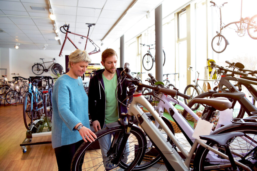 Two people stand indoors in a bicycle shop, looking at a row of modern bikes. The woman in a blue sweater and the man in a dark hoodie appear to be discussing one of the bicycles on display.