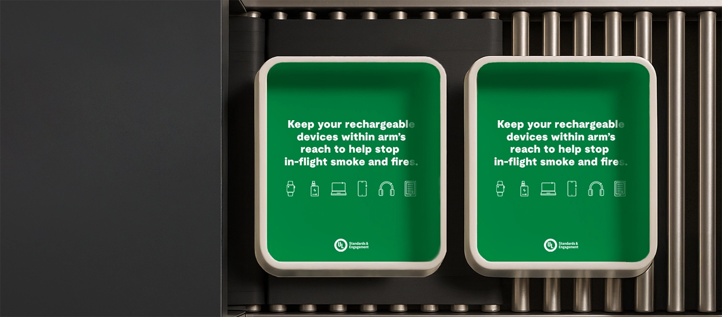 Two green trays on a conveyor belt display aviation safety messages: “Keep your rechargeable devices within arm’s reach to help stop in-flight smoke and fire,” with icons of electronic devices, a safety badge, and reminders about lithium-ion battery fire on planes.