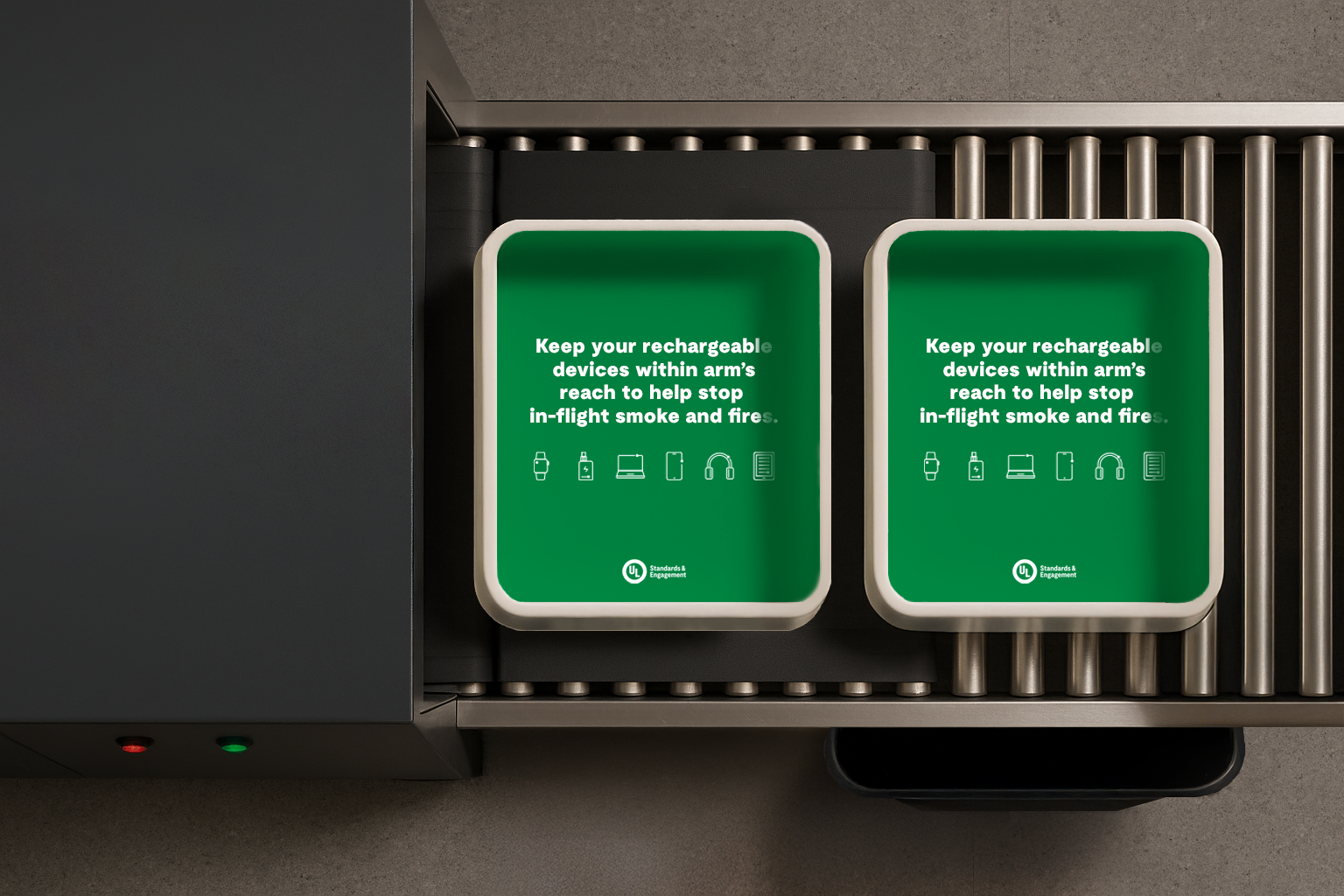 Two green security bins on an airport conveyor belt, each displaying a reminder to keep rechargeable devices within arm’s reach to help prevent lithium-ion battery fire on plane. Simple icons and white text appear on the bins, promoting aviation safety.