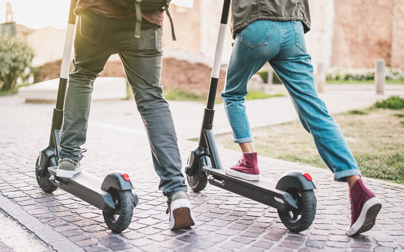 Two people ride electric scooters on a cobblestone path, shown from behind at street level. Both wear casual clothing and sneakers, with the sun shining in the background.