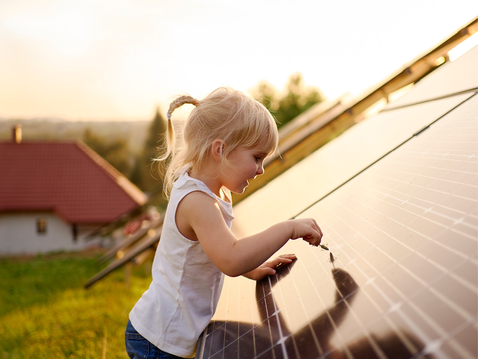 A young child with blonde hair and a white tank top touches the surface of a solar panel outside, with a house and grassy field in the background during sunset.