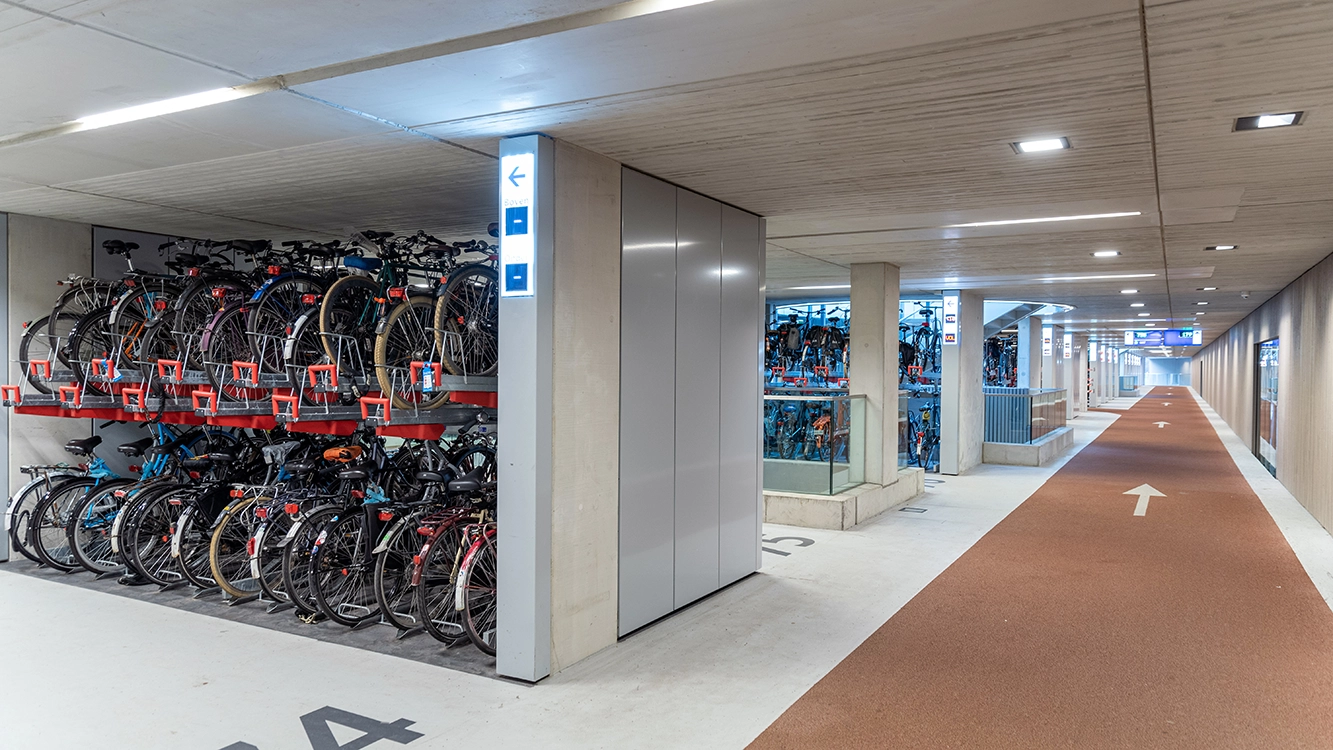 A modern indoor bike parking facility with two-tiered racks holding numerous bicycles, next to a wide, well-lit bike lane with directional arrows on the floor.