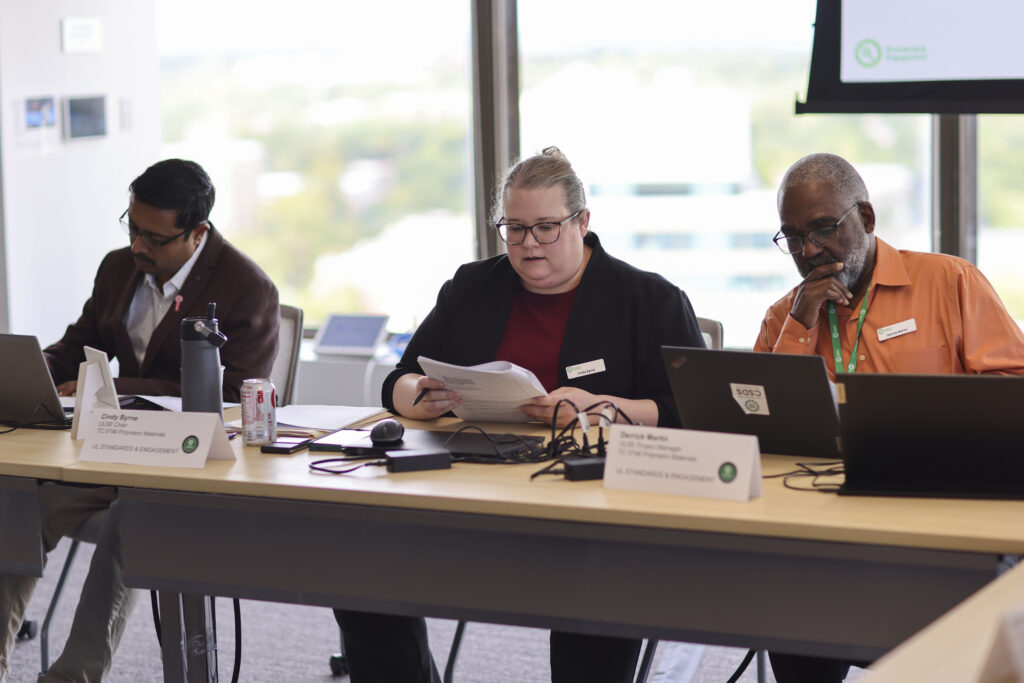Three people sit at a conference table with laptops and documents. The woman in the center, representing UL Standards & Engagement, reads from papers while the two men beside her work on laptops. Name cards and office supplies are visible on the table.