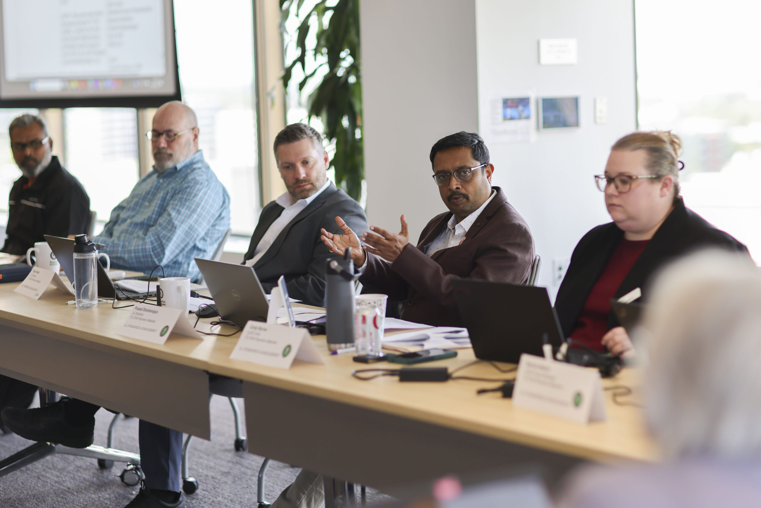 Five people sit at a conference table with laptops and papers, discussing the standards development process. One man gestures as he speaks. Nameplates and water pitchers are on the table; a window and plant are visible in the background.
