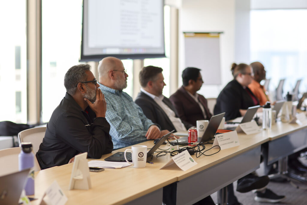 A group of UL Standards & Engagement stakeholders sit at a conference table with laptops, mugs, and nameplates, attentively listening to a presentation on the standards development process in a bright meeting room.