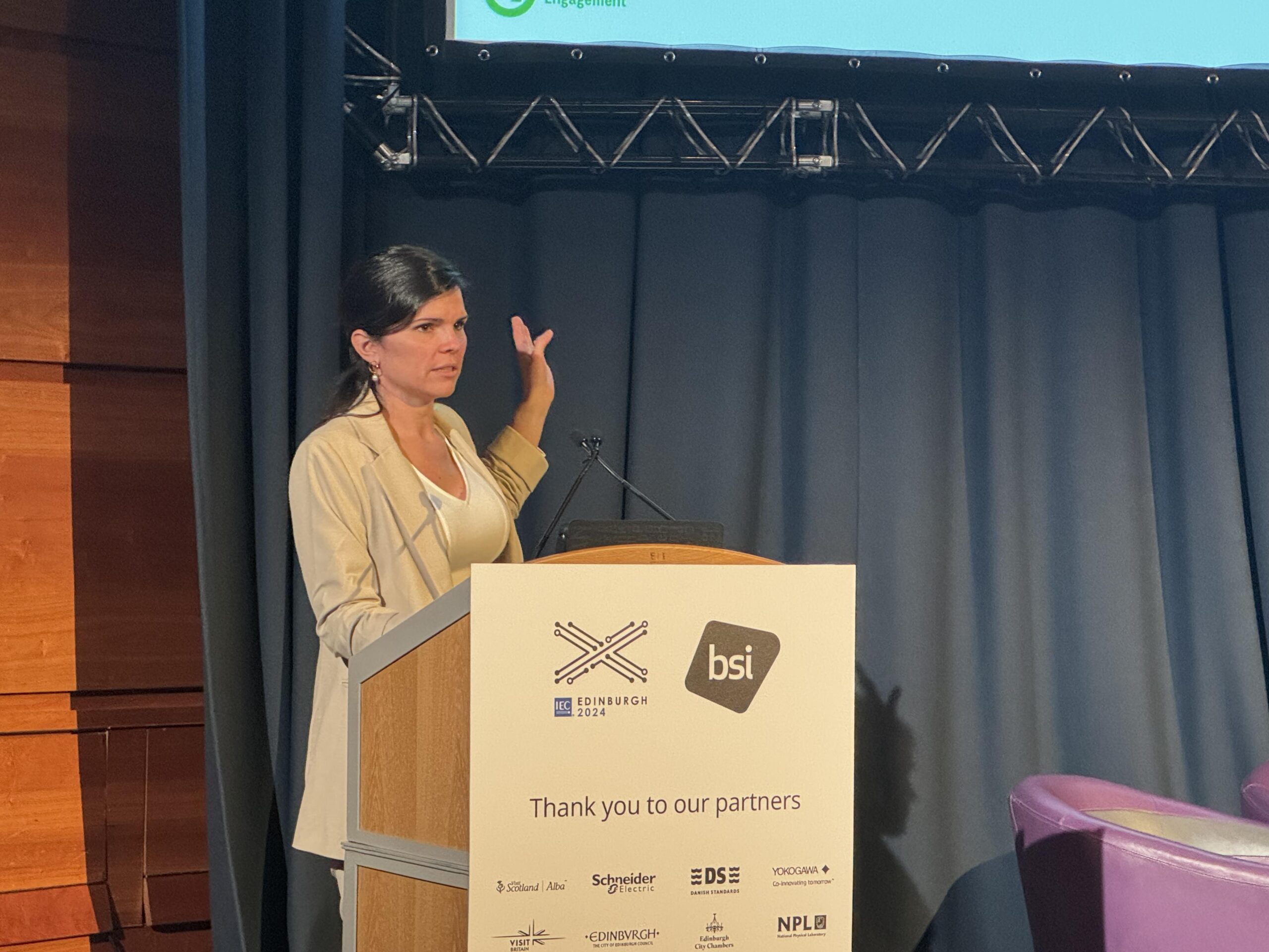 A woman in a beige blazer stands at a podium, gesturing toward a screen while speaking at a conference. The podium displays partner logos, including "bsi," and a sign reads "Thank you to our partners.