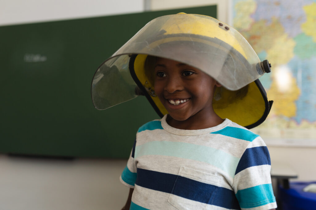 A smiling child wearing a large yellow firefighter’s helmet and a striped t-shirt stands indoors, with a green chalkboard and a colorful map visible in the background.