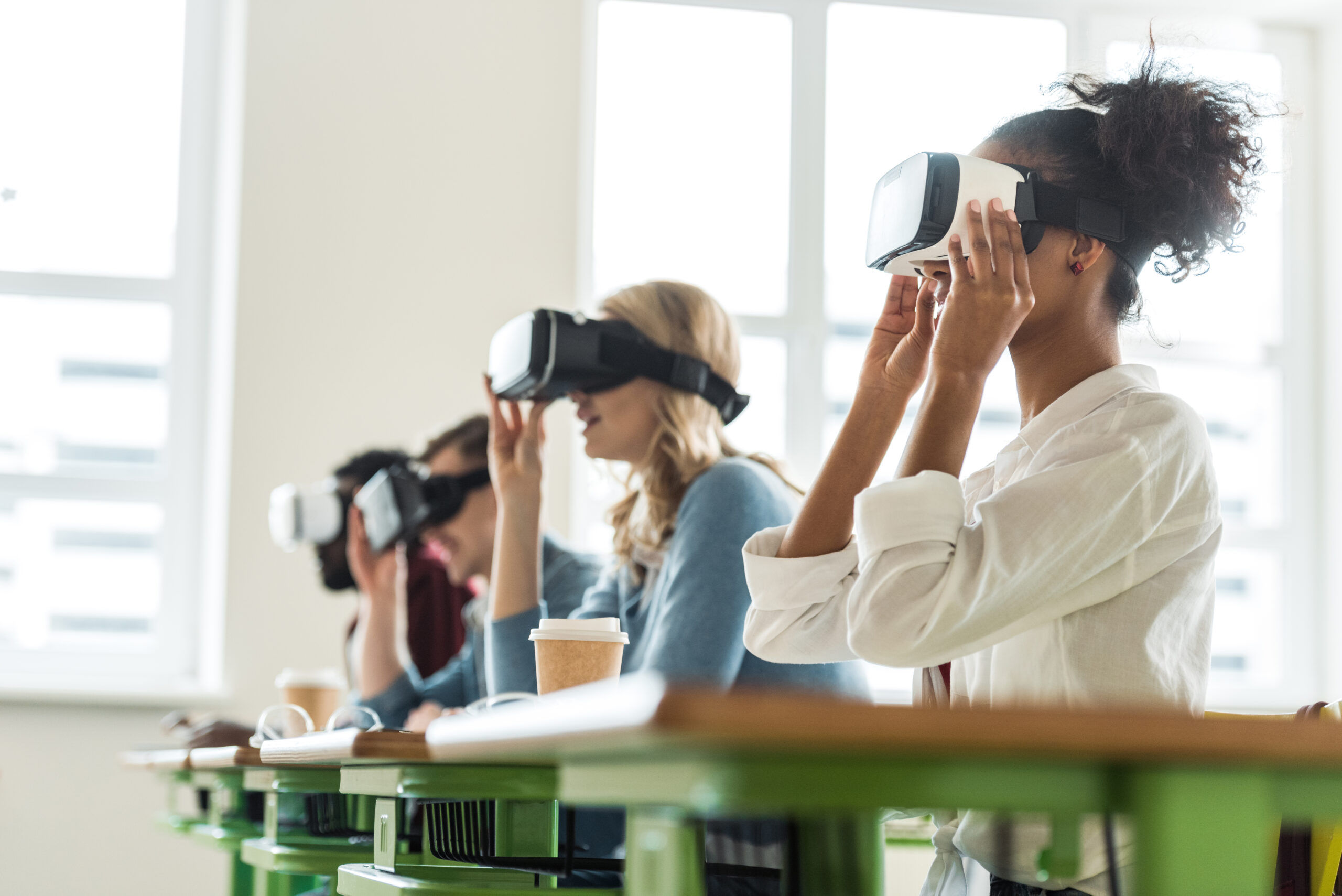 A group of students sitting at desks in a classroom, wearing virtual reality headsets and engaging in an immersive learning experience. Bright daylight streams through large windows in the background.