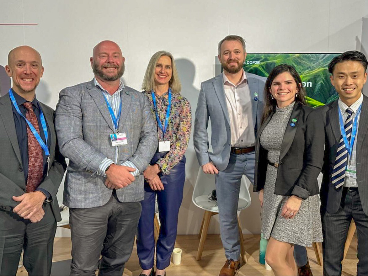 Six people in business attire stand and smile for a group photo indoors, with conference name badges visible. Two chairs and a green display in the background suggest a professional or conference setting.