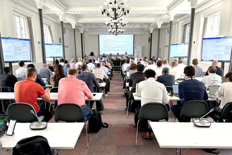 A large conference room filled with people sitting at tables, facing a panel of speakers at the front. Several large screens display information on the side walls. The atmosphere is professional and collaborative.